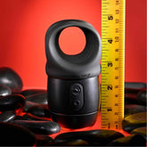 Beside a yellow measuring tape, suggesting exact measurements, the sleek black Zero Tolerance Wet it Be Lube Dispenser Stroker with its rounded loop on top stands prominently. Set against a red backdrop adorned with smooth black stones, this scene sparks curiosity, likely accentuated by the product's enticing multi-speed vibrations.